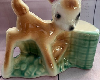 May include: A ceramic planter shaped like a fawn with brown and white fur, pink ears, and black eyes. The fawn is standing on a green and white base.