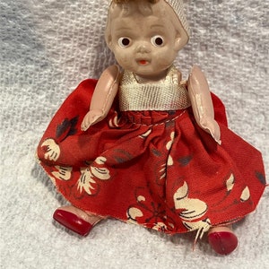 May include: A small porcelain doll wearing a red dress with white floral print and a white bonnet. The doll has brown hair and painted blue eyes.
