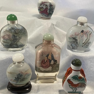 May include: A collection of six antique glass snuff bottles. Each bottle features hand-painted scenes, including landscapes, figures, and floral designs. The bottles have various colored stoppers and are displayed on a white surface.