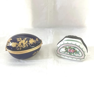 May include: Two decorative porcelain trinket boxes. One is oval-shaped, navy blue with gold floral designs and a gold rim. The other is a half-circle shape with a floral design in pink, green, and white, with a black rim.