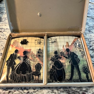 May include: Vintage silhouette art pieces in a box. Each piece features black silhouettes of couples in period clothing, set against a backdrop of a building and a grid pattern. The box is open, revealing the artwork.