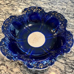 May include: A deep blue ceramic serving bowl with a scalloped edge and gold filigree detailing. The bowl has a central beige circle and radial lines. The rim is white. This decorative bowl is suitable for serving fruit or snacks.