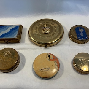 May include: Collection of vintage compacts in various shapes and designs. Includes a square compact with blue wave pattern, a round compact with an anchor, and a compact with the text "SYMPHONIE POWDER".