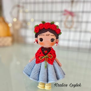 Puede incluir: Una muñeca de crochet que lleva un vestido azul, una chalina roja y una corona de flores. La muñeca tiene el pelo castaño, la cara marrón y ojos negros grandes. La muñeca está de pie sobre una superficie blanca. El texto "Kralice Leylek" es visible en la esquina inferior derecha de la imagen.