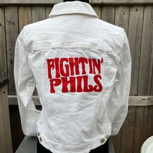 Peut inclure: Veste en jean blanche avec un texte brodé en rouge au dos qui dit "FIGHTIN' PHILS".