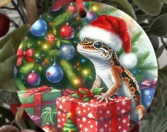May include: A circular Christmas decoration depicting a gecko in a Santa hat, set against a Christmas tree scene. The ornament includes wrapped gifts and baubles, creating a festive holiday theme.