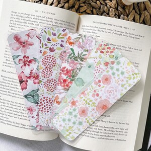 May include: A set of six floral bookmarks with a watercolor design. The bookmarks are made of clear plastic and feature a variety of pink, green, and white flowers. The bookmarks are arranged in a fan shape on top of an open book.