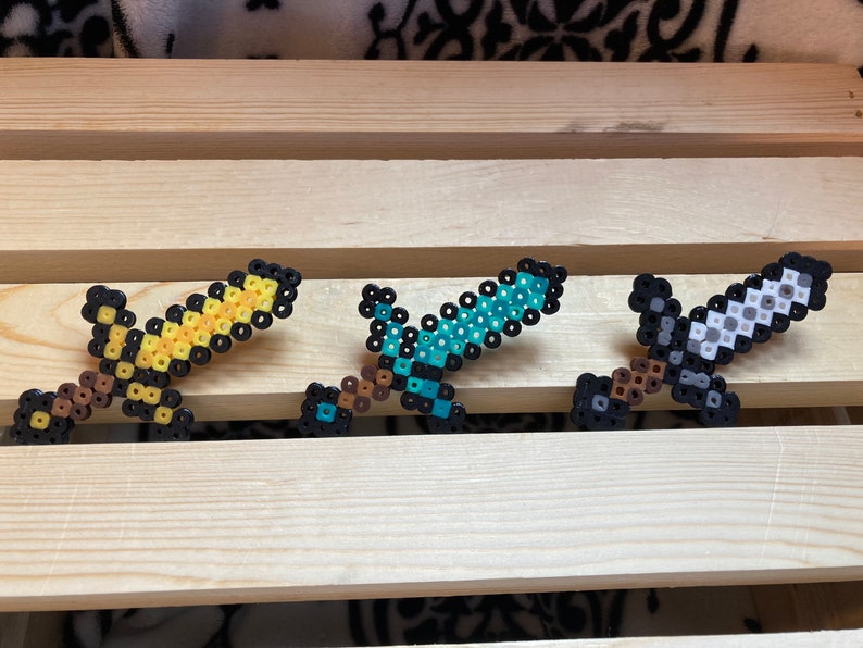 Puede incluir: Tres espadas de pixel art en amarillo, azul y gris, cada una con un mango marr&oacute;n y detalles en negro. Las espadas est&aacute;n hechas de peque&ntilde;as cuentas de colores y se exhiben sobre una superficie de madera.