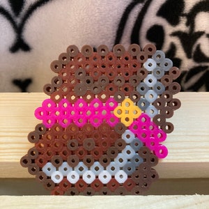 Puede incluir: Un dise&ntilde;o de pixel art de un objeto marr&oacute;n con acentos rosas y amarillos, y una cadena plateada. El objeto est&aacute; hecho de peque&ntilde;as cuentas de colores fusionadas. El dise&ntilde;o est&aacute; sobre una superficie de madera clara.