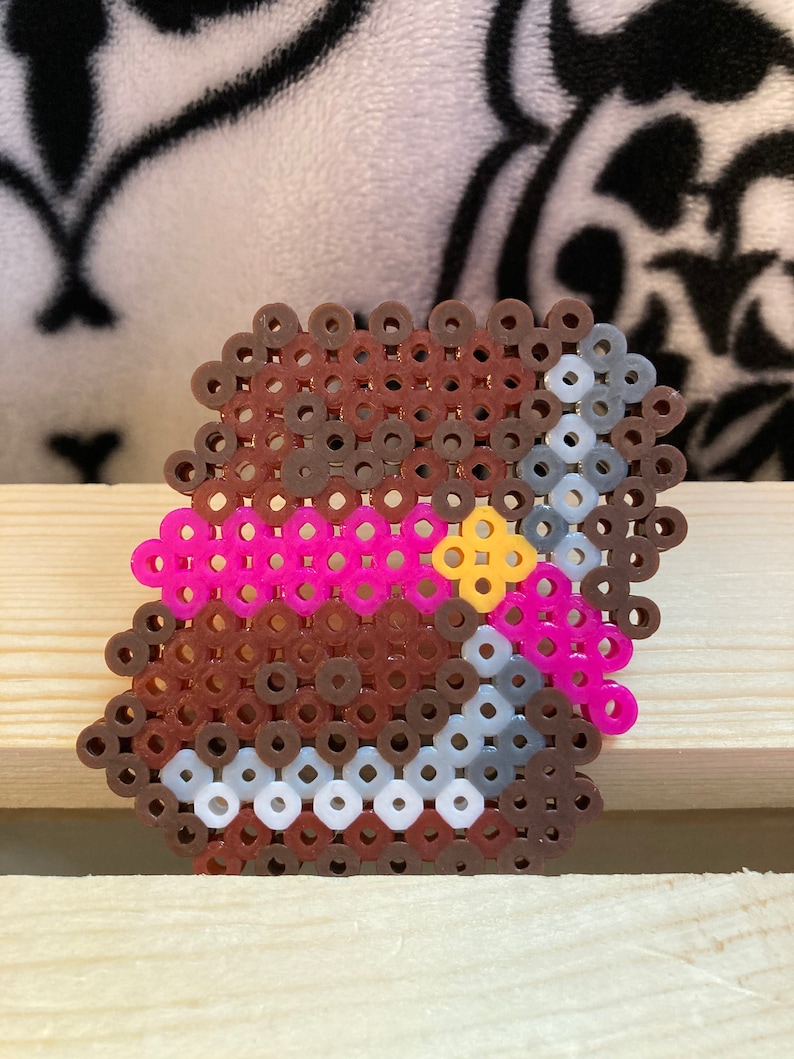 Puede incluir: Un dise&ntilde;o de pixel art de un objeto marr&oacute;n con acentos rosas y amarillos, y una cadena plateada. El objeto est&aacute; hecho de peque&ntilde;as cuentas de colores fusionadas. El dise&ntilde;o est&aacute; sobre una superficie de madera clara.