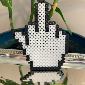 Könnte beinhalten: Eine pixelige Handgeste, hergestellt aus schwarzen und weißen Perlen, die den Mittelfinger zeigt. Das Design ist eine verspielte, Retro-Darstellung eines unhöflichen Handzeichens, perfekt für alle, die einzigartige und ausgefallene Dekorationen mögen.