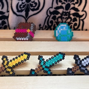 Puede incluir: Creaciones de pixel art que incluyen un cofre marr&oacute;n con una cinta rosa, un diamante azul y verde, y tres espadas. Las espadas son amarillas, azules y grises, todas hechas de cuentas de colores. Los objetos est&aacute;n dispuestos sobre una superficie de madera.