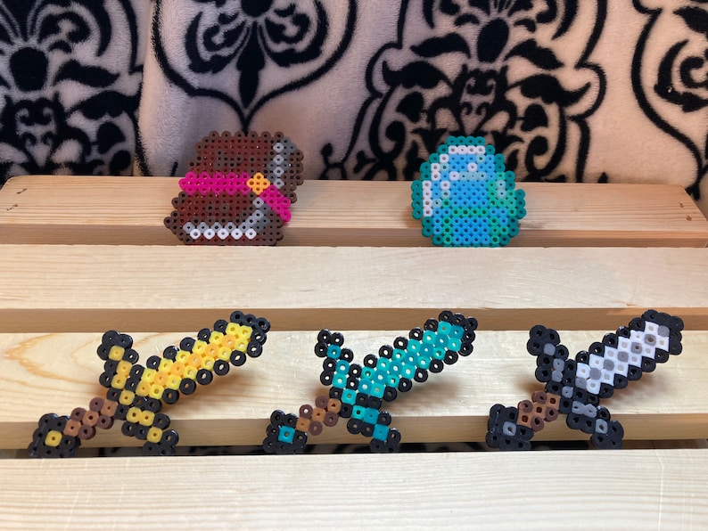Puede incluir: Creaciones de pixel art que incluyen un cofre marr&oacute;n con una cinta rosa, un diamante azul y verde, y tres espadas. Las espadas son amarillas, azules y grises, todas hechas de cuentas de colores. Los objetos est&aacute;n dispuestos sobre una superficie de madera.