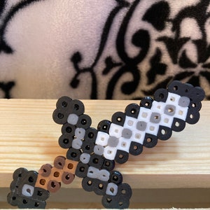 Puede incluir: Una espada de pixel art hecha de peque&ntilde;as cuentas de colores. La espada presenta un mango marr&oacute;n, una hoja gris y blanca y detalles en negro. El art&iacute;culo artesanal se exhibe sobre una superficie de madera.