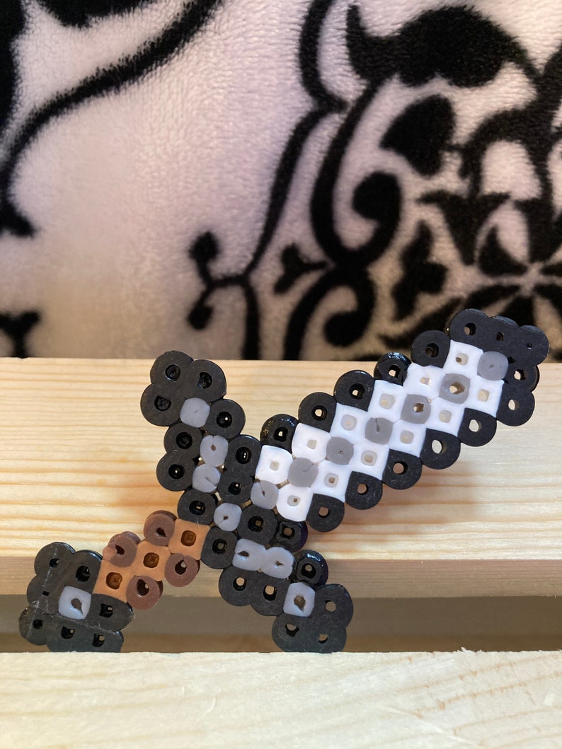 Puede incluir: Una espada de pixel art hecha de peque&ntilde;as cuentas de colores. La espada presenta un mango marr&oacute;n, una hoja gris y blanca y detalles en negro. El art&iacute;culo artesanal se exhibe sobre una superficie de madera.