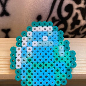 Puede incluir: Un dise&ntilde;o de huevo en pixel art, hecho con peque&ntilde;as cuentas de colores azul, turquesa y blanco. El huevo presenta tonos de azul, turquesa y blanco, dispuestos en un patr&oacute;n de mosaico. El objeto est&aacute; sobre una superficie de madera clara.