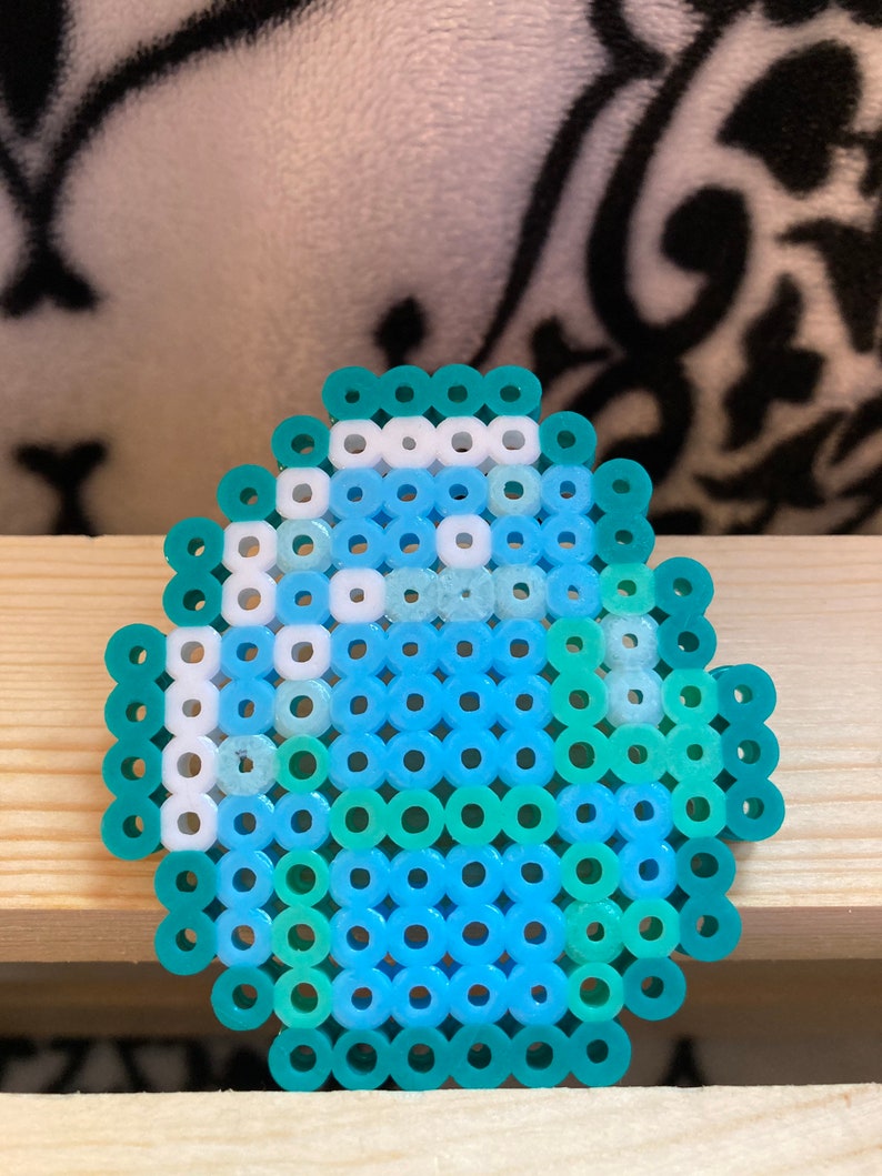 Puede incluir: Un dise&ntilde;o de huevo en pixel art, hecho con peque&ntilde;as cuentas de colores azul, turquesa y blanco. El huevo presenta tonos de azul, turquesa y blanco, dispuestos en un patr&oacute;n de mosaico. El objeto est&aacute; sobre una superficie de madera clara.