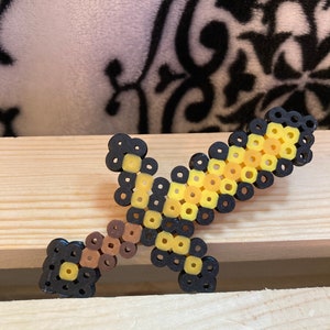 Puede incluir: Una espada pixelada hecha de cuentas de colores. La espada tiene una empu&ntilde;adura y guarda negras, un mango marr&oacute;n y una hoja amarilla y negra. La espada de juguete se exhibe sobre una superficie de madera.