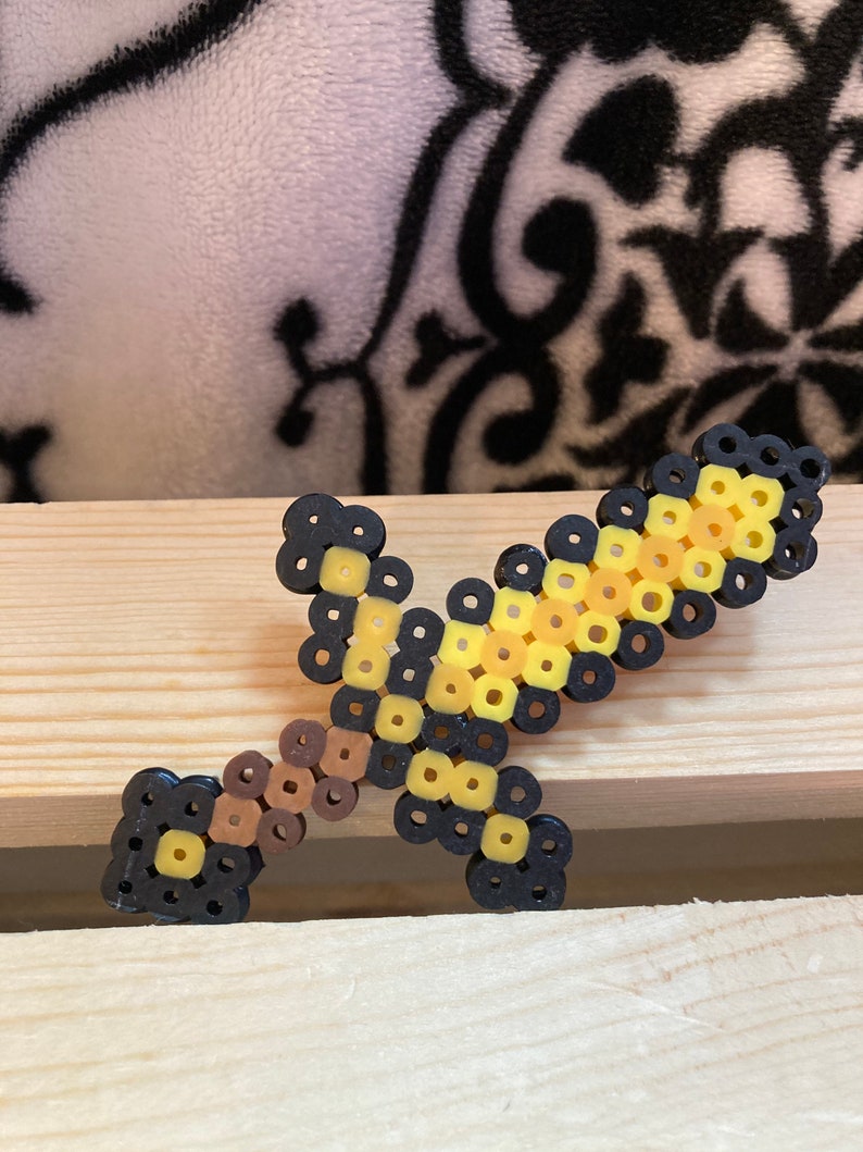 Puede incluir: Una espada pixelada hecha de cuentas de colores. La espada tiene una empu&ntilde;adura y guarda negras, un mango marr&oacute;n y una hoja amarilla y negra. La espada de juguete se exhibe sobre una superficie de madera.