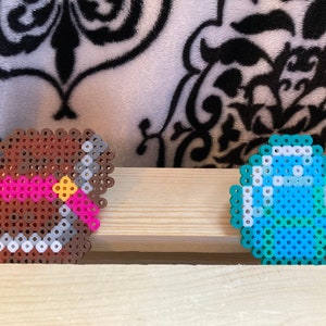 Puede incluir: Dos creaciones de pixel art: un arco y flecha marr&oacute;n con un arco rosa y centro amarillo, y un diamante azul y blanco. Los art&iacute;culos est&aacute;n hechos de peque&ntilde;as cuentas de colores y se colocan sobre una superficie de madera.