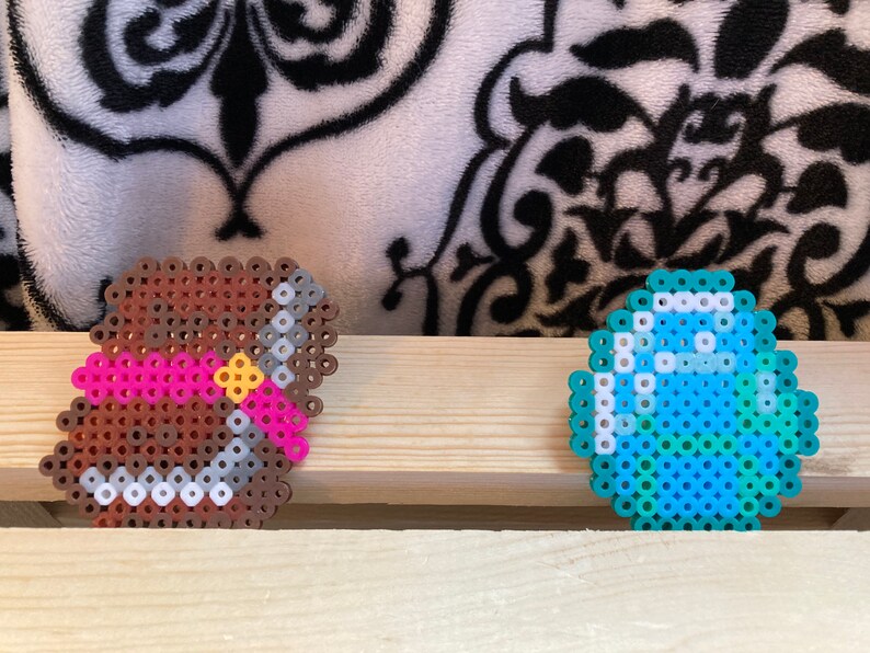 Puede incluir: Dos creaciones de pixel art: un arco y flecha marr&oacute;n con un arco rosa y centro amarillo, y un diamante azul y blanco. Los art&iacute;culos est&aacute;n hechos de peque&ntilde;as cuentas de colores y se colocan sobre una superficie de madera.