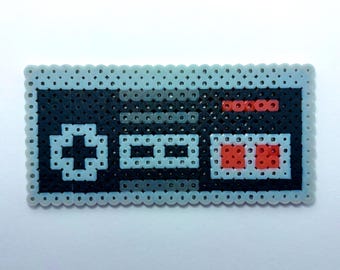 Puede incluir: Una creación rectangular de pixel art hecha de cuentas de plástico fusionadas, diseñada para parecerse a un mando de videojuego clásico. La pieza presenta una base negra con bordes gris claro, una cruceta direccional y dos botones de acción rojos.