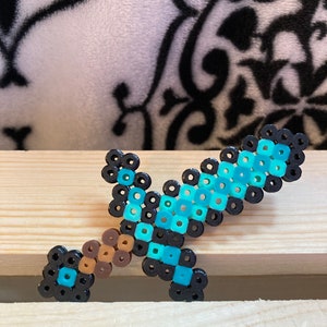 Puede incluir: Una espada pixelada hecha de cuentas de colores. La espada presenta una hoja azul claro, un mango marr&oacute;n y detalles en negro. El art&iacute;culo artesanal se exhibe sobre una superficie de madera.
