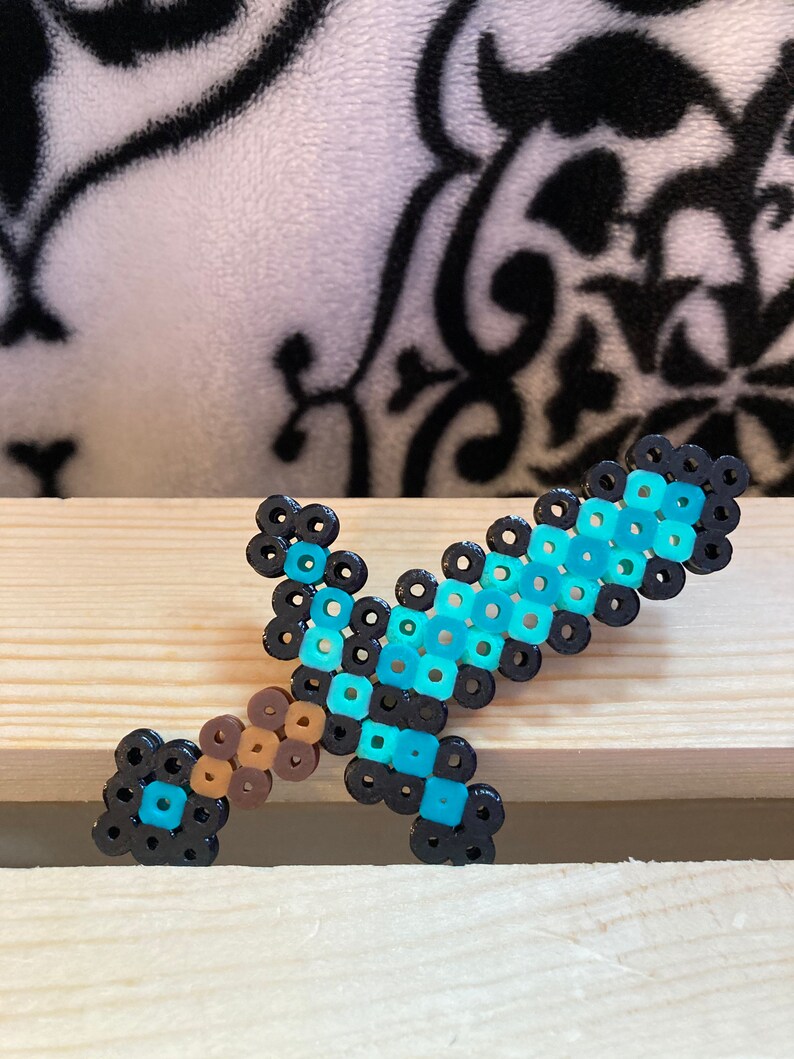 Puede incluir: Una espada pixelada hecha de cuentas de colores. La espada presenta una hoja azul claro, un mango marr&oacute;n y detalles en negro. El art&iacute;culo artesanal se exhibe sobre una superficie de madera.