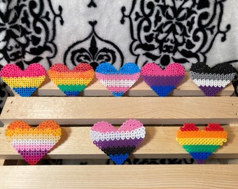 Puede incluir: Nueve creaciones de cuentas en forma de corazón, cada una con un diseño diferente de la bandera del orgullo LGBTQ+. Los corazones están dispuestos sobre una superficie de listones de madera contra un fondo estampado. Los corazones presentan varios colores, incluyendo rosa, azul, amarillo y arcoíris.