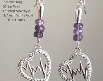 May include: A pair of silver earrings with a heart-shaped pendant featuring a stylized EKG line. The earrings are 2 inches long and feature a 3/4 inch white gold filled heart with amethyst stones.
