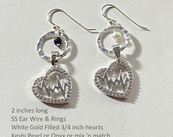 May include: A pair of silver earrings with a heart-shaped pendant featuring a stylized EKG line. Each earring has a hammered silver ring with a pearl or onyx gemstone. The earrings are 2 inches long and the hearts are 3/4 inch.