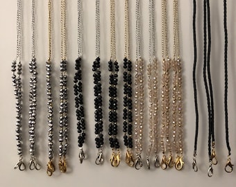 May include: A variety of lanyards for masks, including silver, gold, black, and clear crystal beads, as well as black braided cord. The lanyards are hanging from a white board with the text "Lanyards for Masks".