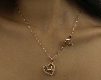 May include: A gold necklace with a heart-shaped pendant featuring a stylized EKG line. The necklace has a toggle clasp with small red beads.