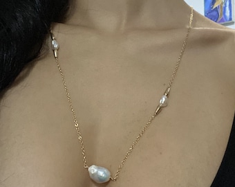 May include: A gold chain necklace with a large, irregular pearl pendant and two smaller pearls. The necklace is on a person's chest.