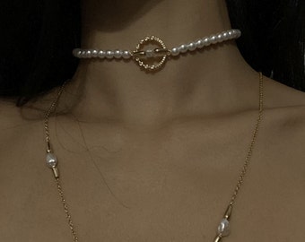 May include: A delicate pearl choker necklace with a gold circle pendant. The necklace is made of small white pearls and has a simple, elegant design.