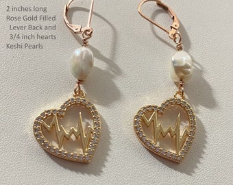 May include: Rose gold filled lever back earrings with a 3/4 inch heart charm featuring a heartbeat design and a white keshi pearl. The earrings are 2 inches long.