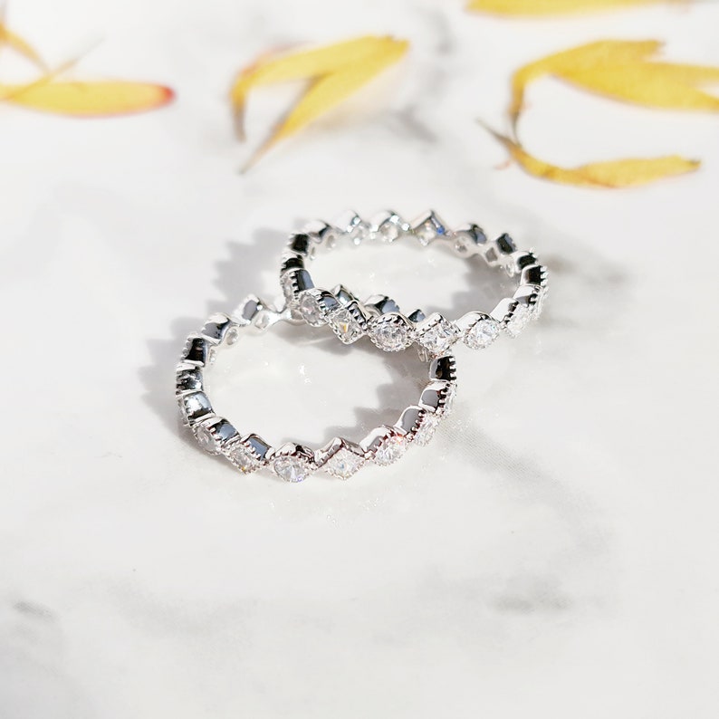 May include: Two silver rings with a row of small, clear, square-shaped stones set in a scalloped design.