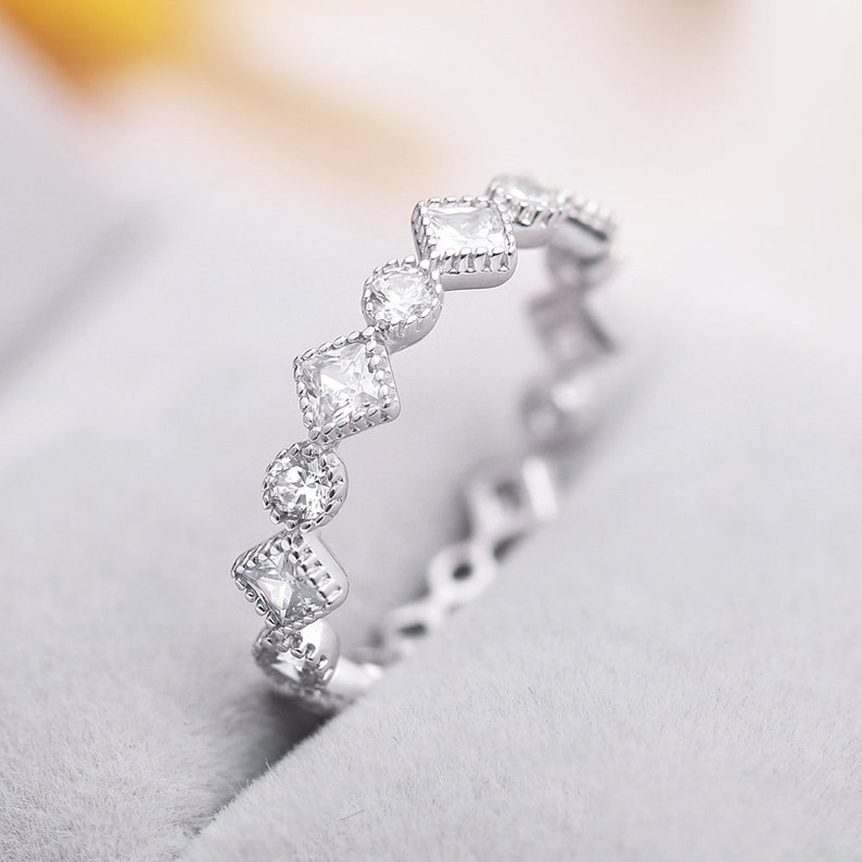 May include: A silver ring featuring a row of small, clear, square-shaped stones set in a delicate, scalloped design.