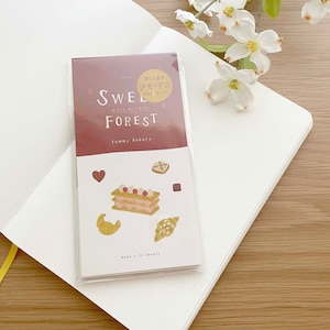 May include: A pink and white notebook with the text "Sweet Forest" and "Yummy Bakery" on the cover. The notebook has a clear plastic cover and is sitting on a wooden surface. The notebook has a small sticker on the front that says "Delicious!  Sweet Choco" in Japanese.