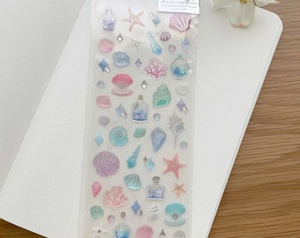 May include: A sheet of clear, colorful,  3D stickers with a seashell theme. The stickers feature seashells, starfish, coral, bottles, and other ocean-themed designs. The sheet is labeled "DROP SEA" and "ST-538".