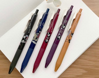 May include: Five black ink pens with Mickey Mouse and Minnie Mouse designs. The pens are in different colors: black, blue, burgundy, purple, and orange. The pens are on a white notebook with a dotted pattern.
