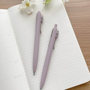 May include: Two lavender Uni-ball One pens lie on an open, lined notebook. The pens have silver clips and the text "Uni-ball One Japan". White flowers and green leaves are in the background, with a wooden surface.