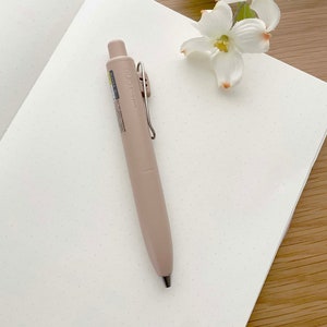 May include: A light pink pen with a silver clip and the word "euno" printed on the side. The pen is lying on a white notebook with small dots.