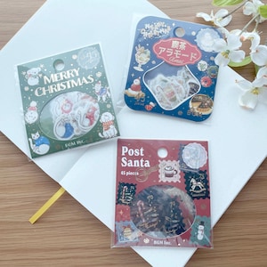 May include: Three packs of Christmas themed stickers. The first pack is green and features a white cat with a red Santa hat. The second pack is blue and features a white cat with a red Santa hat and a cup of coffee. The third pack is red and features a snowman and a rocking horse.
