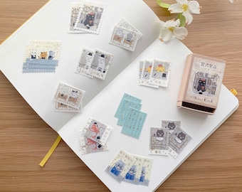 May include: A white notebook with a variety of colorful stickers featuring cats in different poses. The stickers are arranged in rows and columns on the notebook's pages. The notebook is open and lying flat on a wooden surface. A small box with a cat illustration and the text "Shinzi Katoh" is also visible in the image.