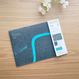 May include: A5 file folder with envelope type pocket. The folder features a gray map design with a turquoise river and text. A white tag with Japanese text is attached. White flowers are in the background.
