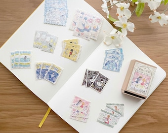 May include: A white notebook with a yellow ribbon bookmark. The notebook is open and has various colorful stickers and stamps on the pages. The stickers and stamps feature illustrations of birds, swans, and people. A small wooden box with a floral design and the text "Shiroi Koibito" is also visible.