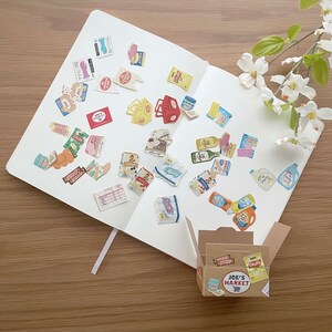 May include: A collection of colorful stickers featuring various grocery items, including food, cleaning supplies, and household goods. The stickers are arranged on a white notebook with a brown cardboard box labeled "Joe's Market" in the bottom right corner.