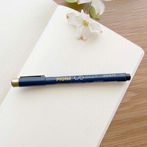 May include: A white notebook with a black and gold pen on top of it. The pen has the text "Pigma 05" on it. The notebook is on a wooden surface.