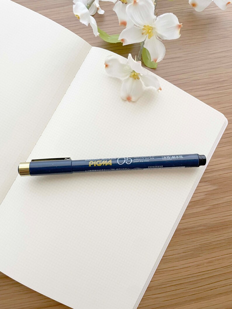 May include: A white notebook with a black and gold pen on top of it. The pen has the text "Pigma 05" on it. The notebook is on a wooden surface.